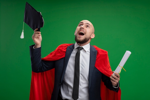 Homme diplômé en cape rouge tenant un diplôme jetant sa casquette heureux et excité debout sur le mur vert