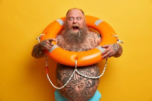 Un homme dodu barbu indigné crie avec colère, pointe directement, a un corps tatoué, pose avec une bouée de sauvetage gonflée, donne l'instuction à nager,