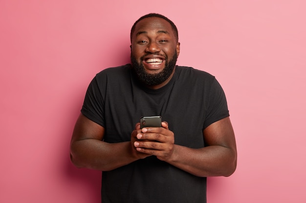 Homme dodu positif avec une barbe épaisse, partage de bonnes nouvelles dans les réseaux sociaux avec un ami, étant sur le nuage neuf du bonheur, tient un smartphone moderne