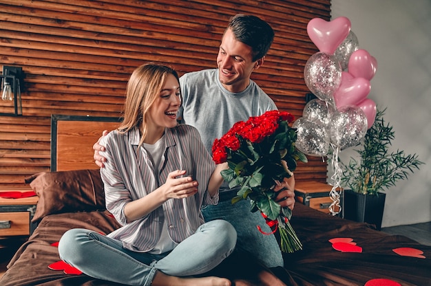 Un homme donne à une femme un bouquet de roses rouges. un couple est assis sur le lit avec des confettis en forme de cœur.