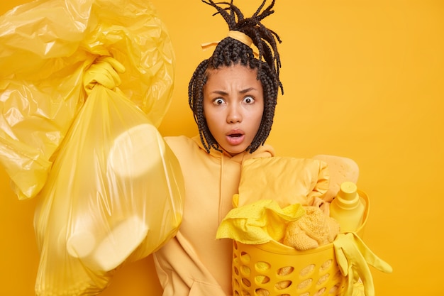 Photo gratuite l'homme avec des dreadlocks peignés porte un sac plein de panier à linge de litière porte un sweat-shirt fait des devoirs domestiques pose sur jaune