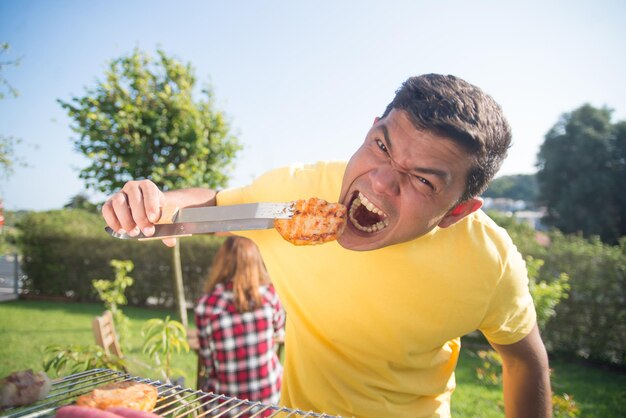 Homme drôle à la fête de famille barbecue. Homme aux cheveux noirs en T-shirt jaune mangeant de la viande fraîchement rôtie et faisant une grimace. BBQ, cuisine, nourriture, concept de famille