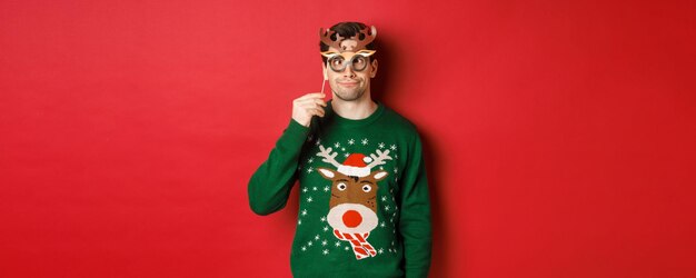 Homme drôle en pull de noël et masque de fête célébrant les vacances d'hiver montrant des grimaces debout ...