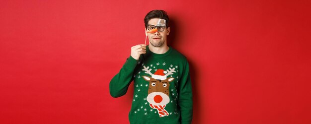 Homme drôle en pull de Noël et masque de fête célébrant les vacances d'hiver montrant des grimaces standi...