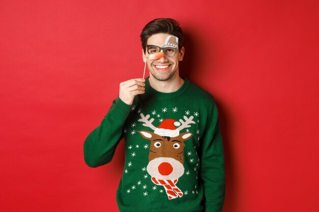 Homme drôle en pull de Noël et masque de fête, célébrant les vacances d'hiver, souriant heureux, debout sur fond rouge