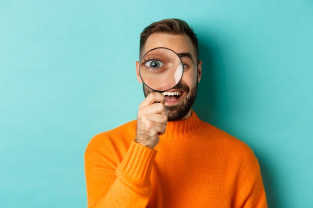 Homme drôle regardant à travers la loupe, la recherche ou l'enquête sur quelque chose, debout dans un pull orange