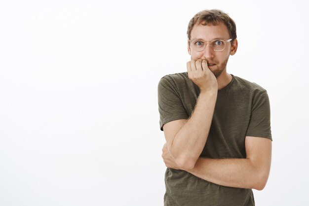 Homme écoutant une rumeur passionnante et intrigante se mordant les ongles de frisson et de stupeur en souriant et en regardant enthousiaste et amusé voulant entendre tous les potins en se tenant debout sur un mur gris