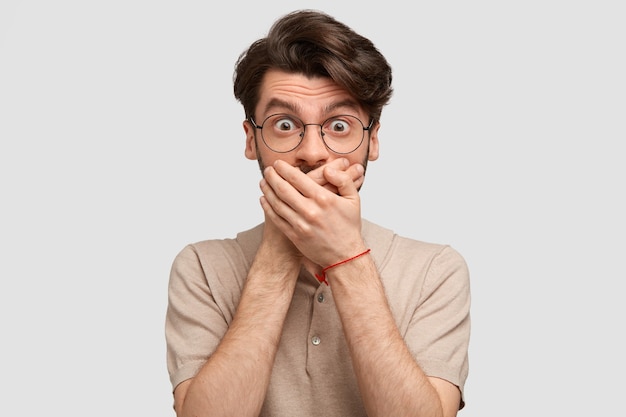 Un homme effrayé débordé avec une coiffure à la mode ferme la bouche avec les mains, essaie d'être muet, se demande quelque chose d'incroyable, vêtu d'un t-shirt beige décontracté