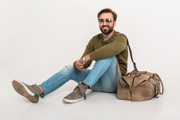 Homme élégant barbu Hansome assis sur le sol isolé habillé en sweat-shirt avec sac de voyage, portant des jeans et des lunettes de soleil