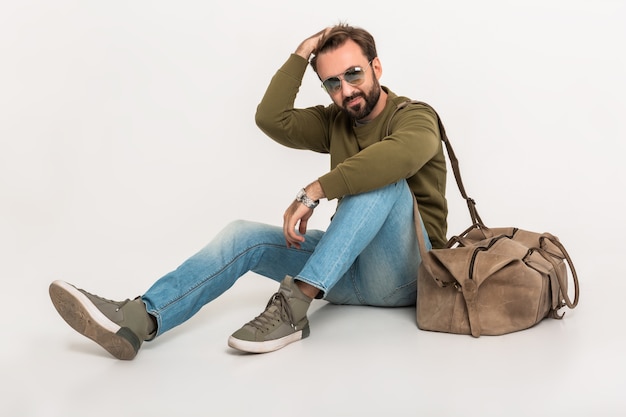 Homme élégant barbu Hansome assis sur le sol isolé habillé en sweat-shirt avec sac de voyage, portant des jeans et des lunettes de soleil