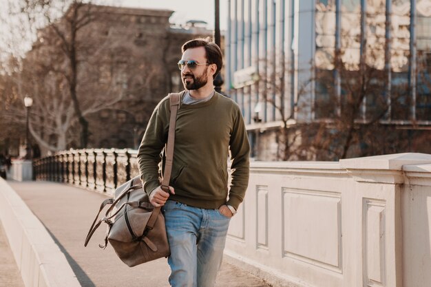 Homme élégant hipster attrayant marchant dans la rue de la ville avec sac en cuir portant sweatshot et lunettes de soleil, tendance de style urbain, journée ensoleillée