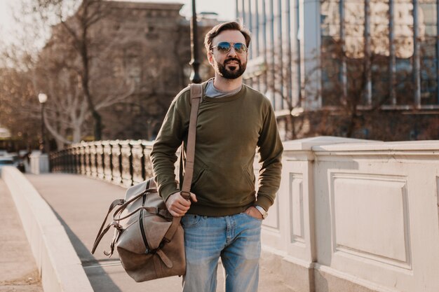 Homme élégant hipster attrayant marchant dans la rue de la ville avec sac en cuir portant sweatshot et lunettes de soleil, tendance de style urbain, journée ensoleillée