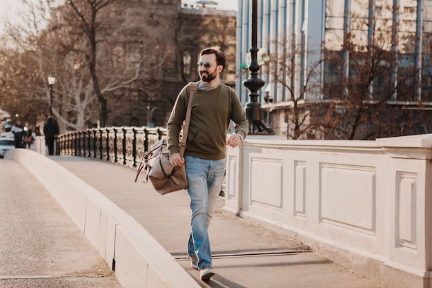 Homme élégant hipster attrayant marchant dans la rue de la ville avec sac en cuir portant sweatshot et lunettes de soleil, tendance de style urbain, journée ensoleillée