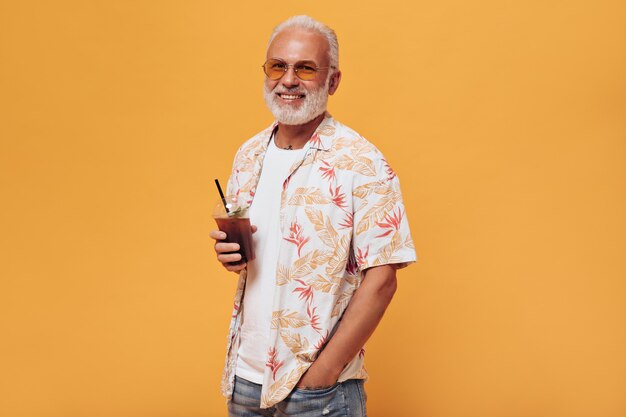 Un homme élégant à lunettes tient un cocktail pour aller sur un mur orange