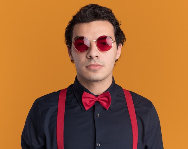 Homme élégant avec noeud papillon portant des lunettes et des bretelles à l'avant avec une expression confiante sérieuse debout sur un mur orange