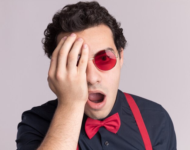 Homme élégant avec noeud papillon portant des lunettes et des bretelles à l'avant surpris couvrant un œil avec bras debout sur un mur blanc