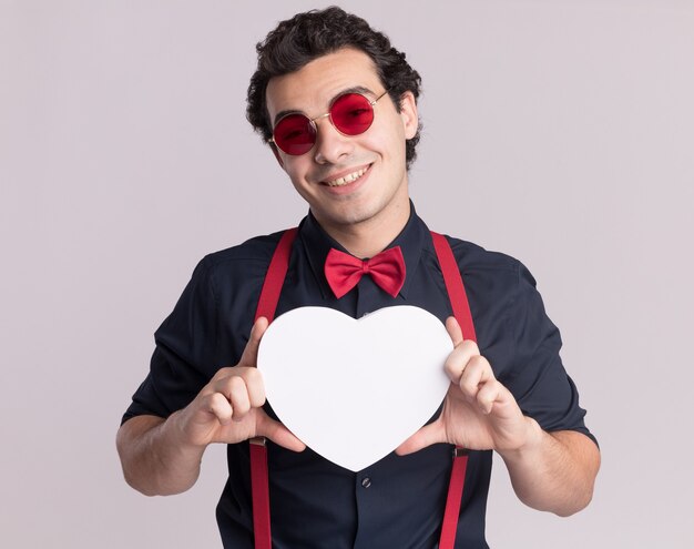 Homme élégant avec noeud papillon portant des lunettes et des bretelles tenant un coeur en carton à l'avant souriant joyeusement debout sur un mur blanc