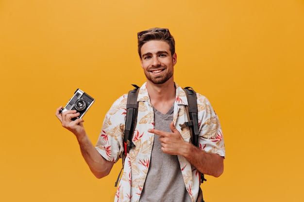 Homme élégant positif en chemise légère à la mode et t-shirt gris posant avec caméra et souriant sur fond isolé orange
