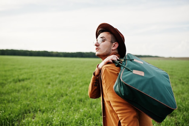 Homme élégant en veste marron lunettes et chapeau avec sac posé sur champ vert