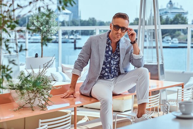 Un homme élégant vêtu de vêtements modernes et élégants corrige ses lunettes de soleil tout en étant assis sur une table au café en plein air sur fond de quai de la ville.
