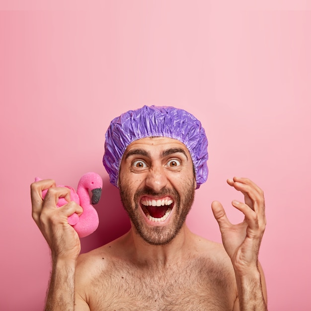 L'homme émotif fait des gestes actifs, étant agacé, tient une éponge flamant rose tendre, porte un bonnet de douche