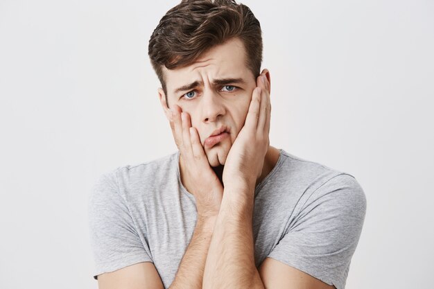 Un homme émotionnel stupéfait choqué garde les mains sur les joues, étant troublé d'écouter les conseils de ses parents isolés sur un fond de mur de studio vide. Un homme européen ne peut pas croire aux nouvelles choquantes