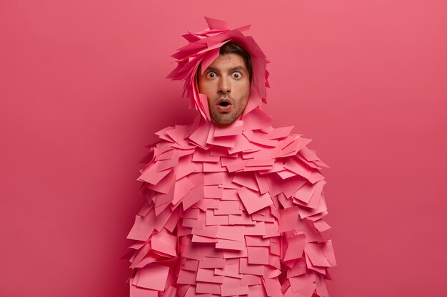 Un homme émotionnel stupéfait regarde avec des yeux écarquillés, exprime un choc, porte des vêtements créatifs faits de notes autocollantes, ouvre la bouche avec émerveillement, isolé sur un mur rose, entend les dernières nouvelles, de mauvaises conceptions