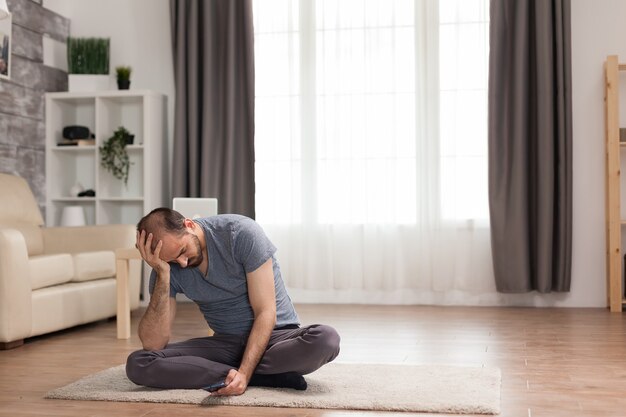 Homme ennuyé assis sur un tapis dans le salon naviguant sur un smartphone pendant la quarantaine.