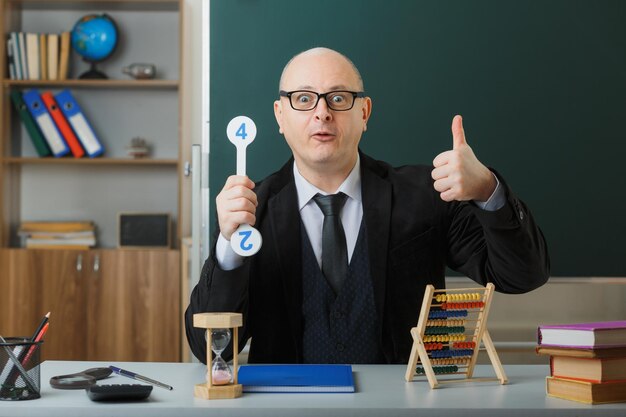 Homme enseignant portant des lunettes assis au bureau de l'école avec registre de classe devant le tableau noir dans la salle de classe tenant des plaques d'immatriculation expliquant la leçon montrant le pouce vers le haut