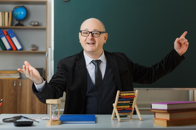 Homme enseignant portant des lunettes avec registre de classe assis au bureau de l'école devant le tableau noir dans la salle de classe heureux et positif levant le bras pointant avec l'index sur le tableau noir