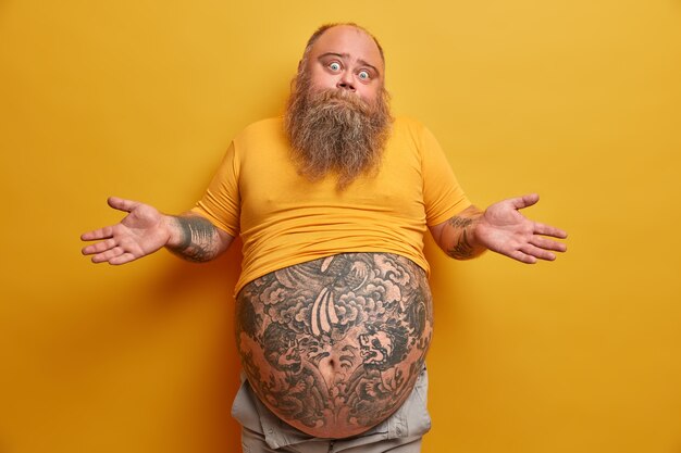 Homme épais hésitant avec un gros ventre tatoué, hausse les épaules et a l'air confus, fait face à un dilemme, prend une décision sérieuse, porte un t-shirt jaune sous-dimensionné, pose à l'intérieur. Concept de personnes et de doute