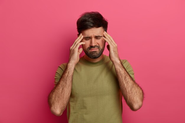 Un homme épuisé touche les tempes avec les yeux fermés, souffre de maux de tête, attend que quelqu'un apporte des analgésiques, porte un t-shirt, passe une mauvaise journée, isolé sur un mur rose, dérangé par une maladie douloureuse