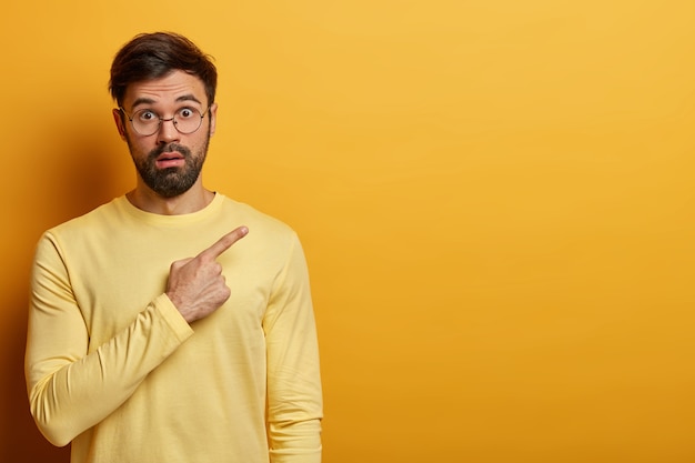 Un homme étonné stupéfait annonce une présentation incroyable, pointe sur l'espace de copie vierge du coin supérieur droit, halète d'émerveillement, porte un pull jaune sur un ton avec un mur, fait la publicité du produit