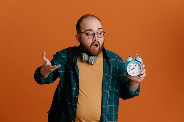 Homme étudiant en tenue décontractée portant des lunettes avec un casque tenant un réveil le regardant inquiet et confus debout sur fond orange