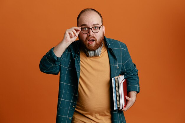 Homme étudiant en vêtements décontractés portant des lunettes avec des écouteurs tenant des cahiers regardant la caméra surpris et étonné debout sur fond orange