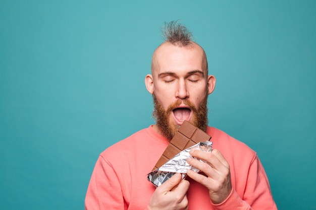 Homme européen barbu en pêche occasionnel isolé, appréciant le chocolat avec les yeux fermés morceau de morsure