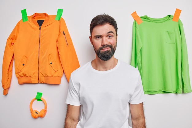 Photo gratuite un homme européen sérieux et hésitant porte les lèvres à l'air indigné par la caméra porte un t-shirt décontracté pose à l'intérieur sur fond blanc avec un pull en anorak plâtré et des écouteurs pense quoi s'habiller