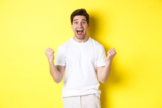 Homme excité et chanceux qui gagne, serre les poings et a l'air heureux, triomphant et célébrant, debout sur fond jaune.