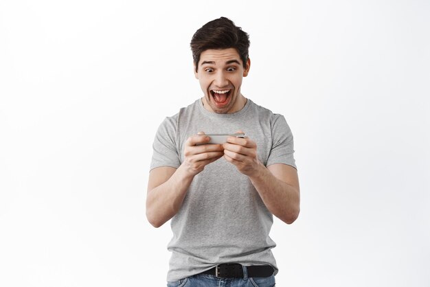 Homme excité jouant au jeu vidéo sur smartphone et regardant joyeux à l'écran souriant étonné Guy regardant le flux en direct en ligne sur l'application de téléphone debout sur fond blanc