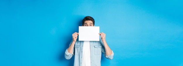 Homme excité se cachant le visage derrière une place de papier vierge pour votre logo debout sur fond bleu