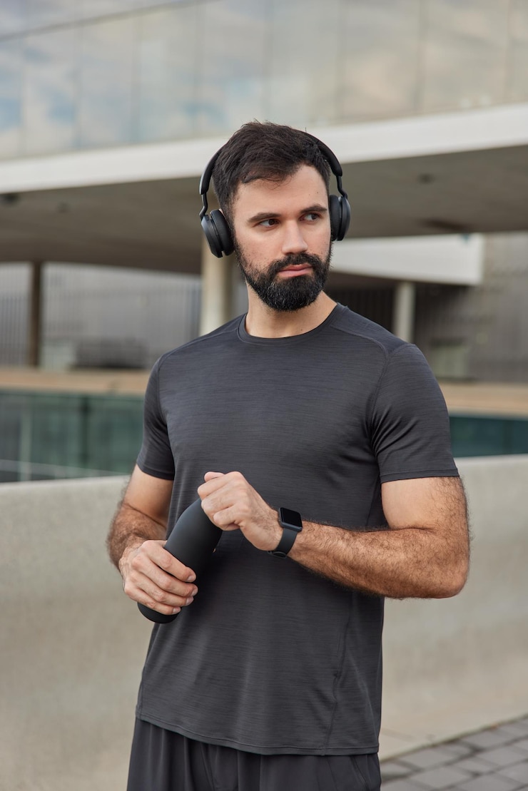 Photo gratuit l'homme fait du sport prend une pause écoute de la musique de motivation dans des écouteurs sans fil se rafraîchit avec de l'eau porte des poses de t-shirt décontractées sur des exercices flous à l'extérieur