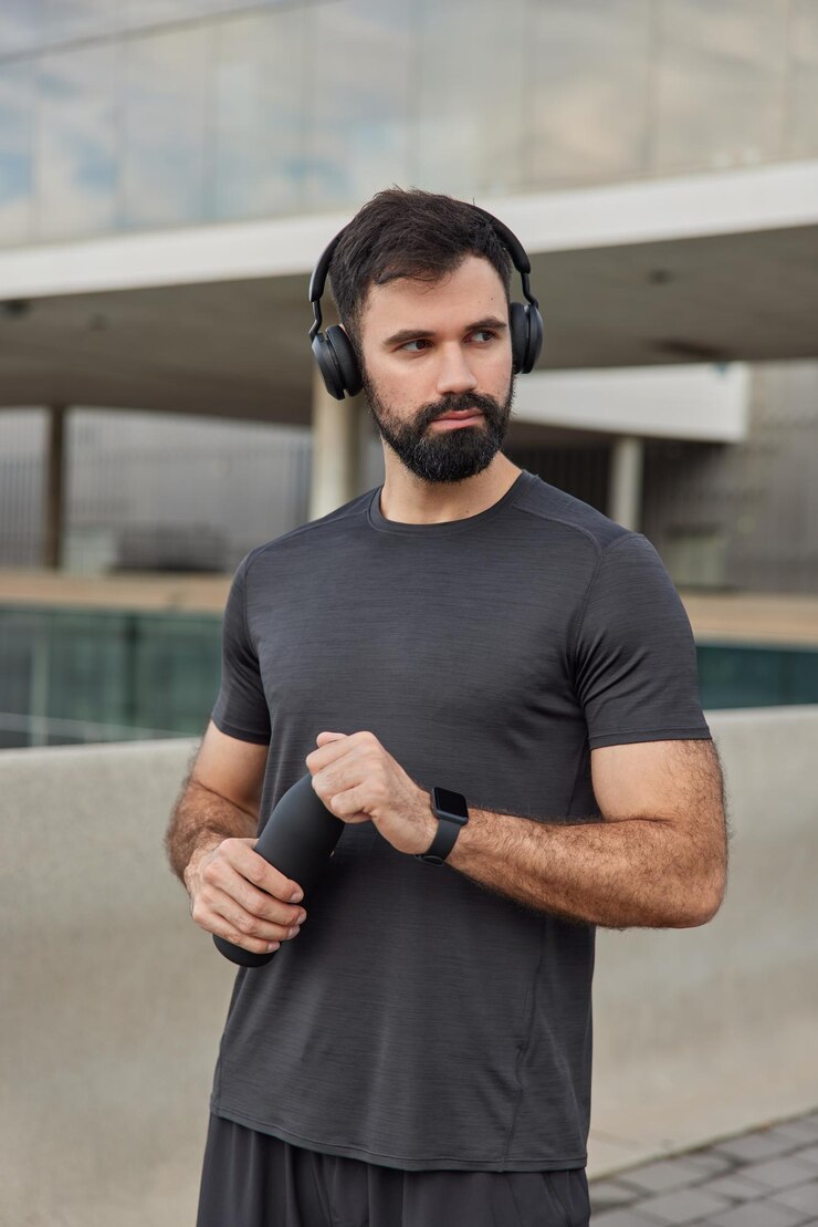 Photo gratuit l'homme fait du sport prend une pause écoute de la musique de motivation dans des écouteurs sans fil se rafraîchit avec de l'eau porte des poses de t-shirt décontractées sur des exercices flous à l'extérieur