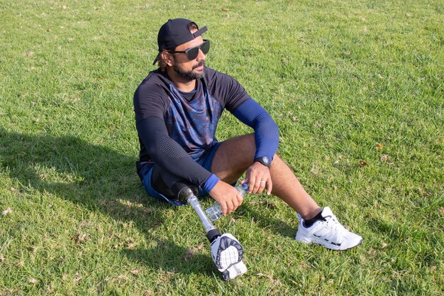 Homme fatigué avec une jambe prothétique se relaxant après l'entraînement. Homme caucasien avec barbe tenant de l'eau en bouteille, assis sur l'herbe. Concept sport, loisirs, handicap