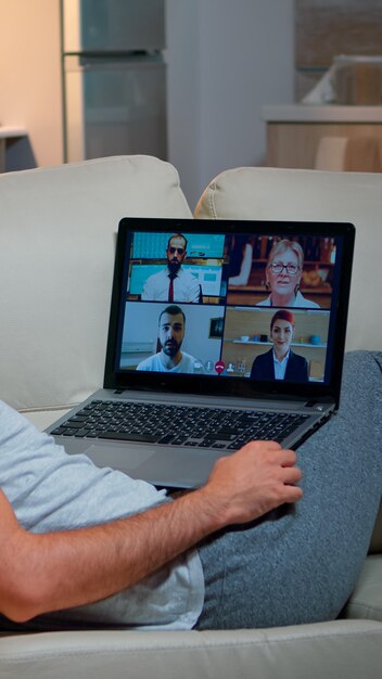 Homme fatigué en pyjama assis sur un canapé s'endormant tout en ayant un appel vidéo d'affaires en ligne avec des coéquipiers à l'aide d'un ordinateur portable. Homme de race blanche sur la communication web de la conférence internet