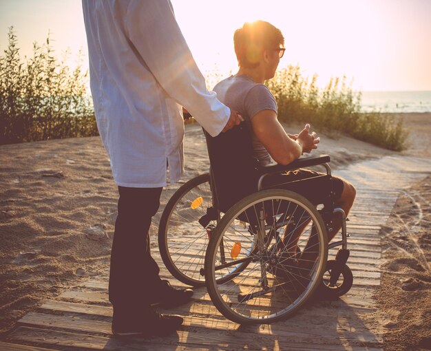 Un homme en fauteuil roulant et son infirmière profitant du lever du soleil sur une plage.