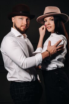 Un homme et une femme en chemises blanches et chapeaux sur fond noir. un couple amoureux pose à l'intérieur du studio.