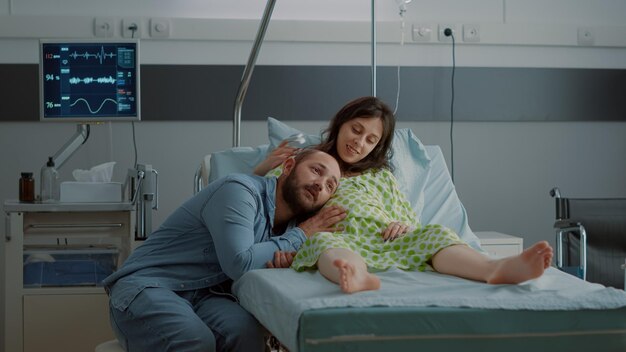 Un homme et une femme de race blanche attendent un enfant dans une salle d'hôpital tandis qu'une infirmière afro-américaine répare un sac d'égouttement IV pour une mère enceinte à la maternité. Jeune couple heureux en attente d'accouchement