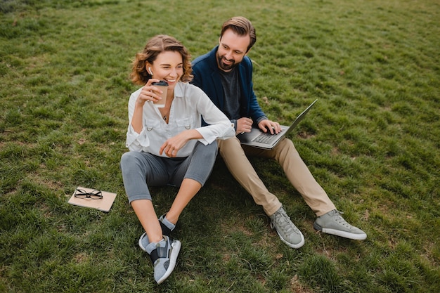 Homme et femme souriants séduisants parlant assis sur l'herbe dans un parc urbain, prenant des notes