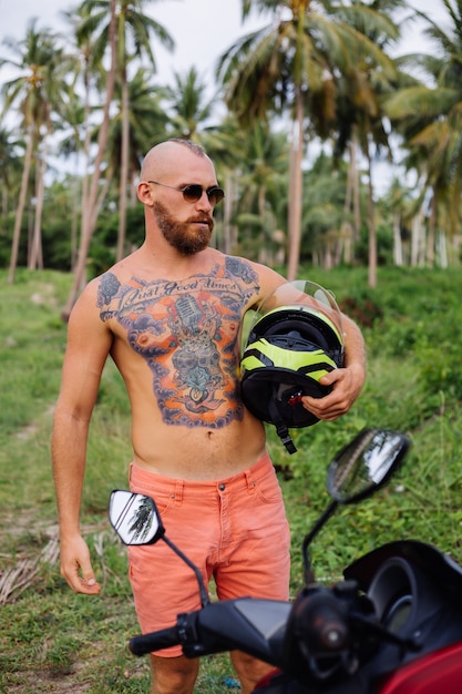 Homme fort tatoué sur champ de jungle tropicale avec moto rouge