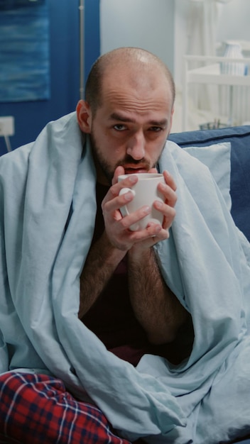 Photo gratuite homme avec des frissons froids et buvant une tasse de thé tout en étant enveloppé dans une couverture. personne malade tenant une tasse avec une boisson chaude contre les frissons et les symptômes de fièvre tout en regardant la caméra. adulte grippé